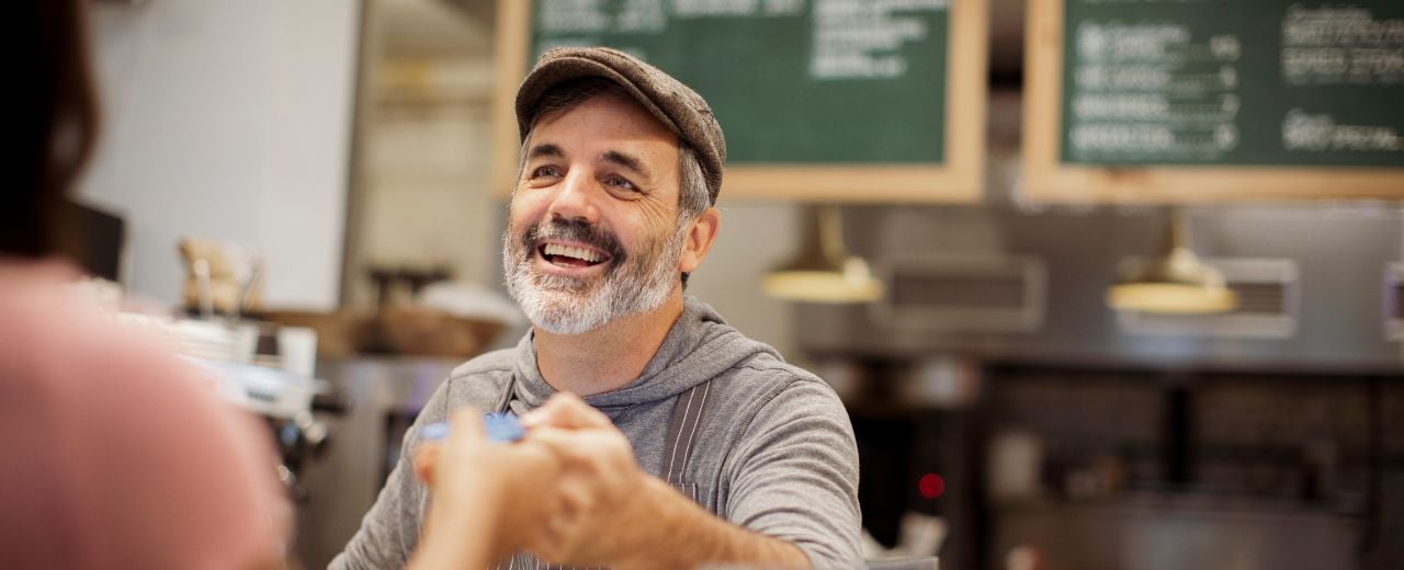 At the cash register, the café owner smiles while giving great service to a customer.